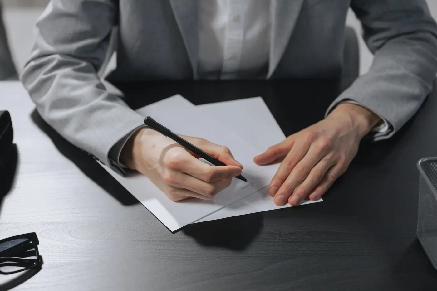 A person in a grey suit signing a document at a dark desk