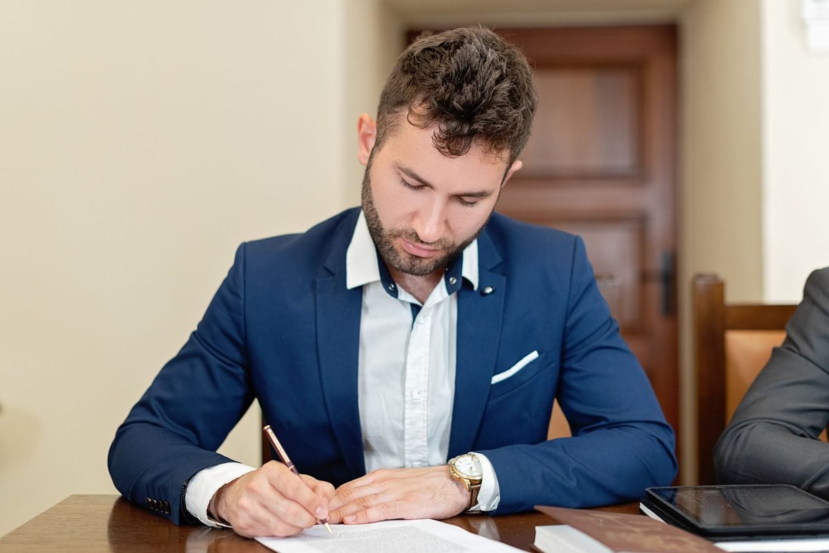 Man in a blue jacket signing a contract at a wooden office desk